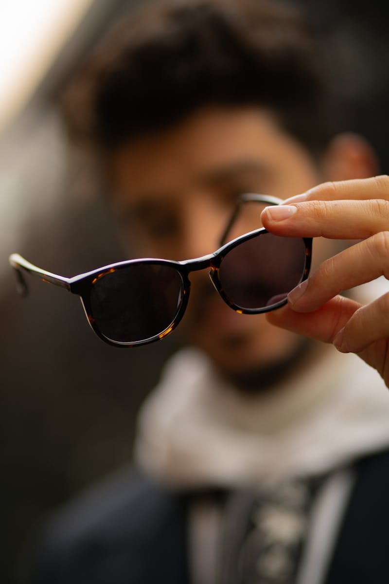 Stylish man holding sunglasses with blurred background, emphasizing fashion accessories.